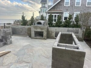 Early stage of a stone outdoor kitchen installation showing masonry layout for pizza oven, grill, and prep area on a flagstone patio.