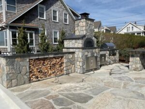 Full façade of finished outdoor kitchen featuring pizza oven, dual cabinets, and stacked firewood.